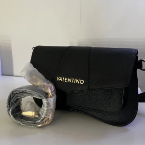 Svart axelväska från Valentino - Stilren svart axelväska från Valentino med guldfärgad logga framtill. Väskan har en elegant, böljande form och justerbar axelrem med gulddetaljer. Tillverkad i ett strukturerat syntetmaterial som ger en lyxig känsla. Perfekt för dig som vill ha en snygg och trendig accessoar. Kommer med en kort och en lång rem. (Den korta är oöppnad och fortfarande i sin orginalförpakning)