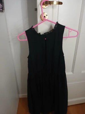 Svart ärmlös klänning från bpc - Svart ärmlös klänning från bpc med volangdetalj vid halsen och dekorativa pärlor i midjan. Klänningen har en luftig och lätt form, perfekt för dig som gillar stilrena och enkla plagg med en touch av glitter. Materialet känns som polyester och har ett skirt, snyggt fall.