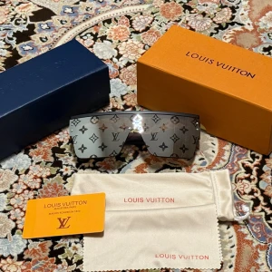 Louis Vuitton monogram solglasögon - Snygga och futuristiska solglasögon från Louis Vuitton med spegelglas och ikoniskt monogrammönster över hela linsen. Sköldformad design med svart båge och silverfärgade glas. Kommer med originalbox, fodral och certifikat.