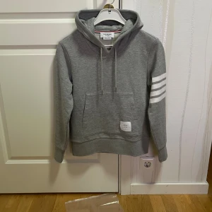 Thom Browne. Hoodie - Snygg grå hoodie från Thom Browne med klassiska vita ränder på ena ärmen och en patch med logga vid fickan. Tröjan har huva med snörning, stor magficka och ribbade muddar. Perfekt för dig som gillar stilren streetwear.