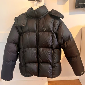 Svart pufferjacka från Calvin Klein Jeans - Svart pufferjacka från Calvin Klein Jeans med hög krage och dragkedja framtill. Jackan har två sidofickor med dragkedja, broderad CK-logga på bröstet och Calvin Klein-tryck på ärmen. Materialet är glansigt och syntetiskt, perfekt för kalla dagar.