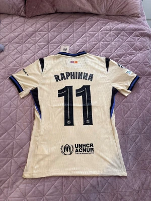 Barcelona Raphinha bortatröja Nike - Snygg beige Barcelona bortatröja från Nike med Raphinha 11 på ryggen. Tröjan har svarta detaljer, klubbmärke, sponsorloggor och blåsvarta ränder vid krage och ärmslut. Tillverkad i lätt funktionsmaterial, perfekt för fotboll eller streetwear.