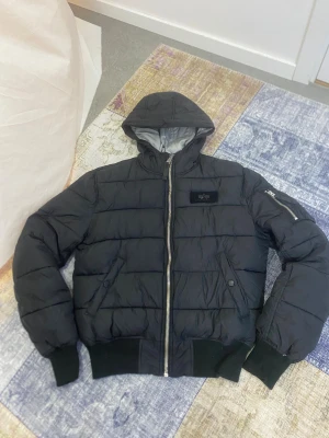 Svart pufferjacka Alpha Industries S - Svart pufferjacka från Alpha Industries i storlek S. Jackan har huva, silverfärgad dragkedja, ribbade muddar och flera fickor, inklusive en med dragkedja på ärmen. Insidan är ljusgrå och materialet är syntet. Perfekt för kalla dagar och har en clean streetstil.