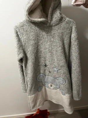 Grå fluffig hoodie med nalle från KappAhl - Supermjuk och fluffig hoodie från KappAhl i grått och vitt, med gulligt nallemotiv och stjärnor framtill. Tröjan har huva och långa ärmar, perfekt för att mysa hemma. Materialet är fleece och ger en skön känsla mot huden.