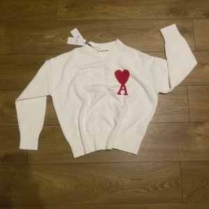 Vit stickad tröja från Ami Paris - Snygg vit stickad tröja från Ami Paris med röd hjärtapplikation och bokstaven A på bröstet. Tröjan har rund hals, ribbade muddar och långärmad passform. Perfekt för dig som gillar stilrena detaljer och vill ha något som sticker ut.