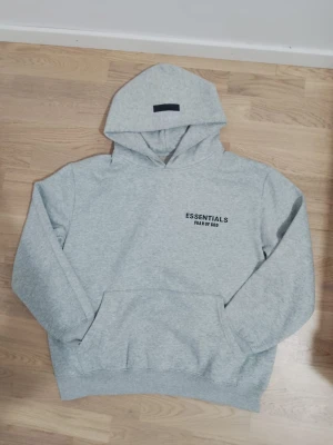 Grå Essentials Fear of God hoodie - Snygg ljusgrå hoodie från Essentials Fear of God med svart logotyptryck både fram och bak. Tröjan har huva, känguruficka och ribbade muddar. Perfekt för dig som gillar streetwear och vill ha en clean look. Materialet är mjuk bomullsmix.