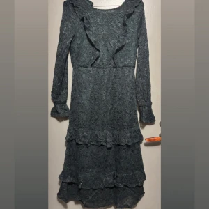 Mörk grön spetsklänning från Lindex - Klänning med spetsmaterial och volangdetaljer både upptill och nedtill. Klänningen har långärmade ärmar med volang vid mudden och en vacker öppen rygg. 