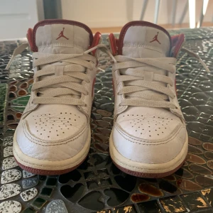 Nike Air Jordan 1 Mid vit/röd US 13C - Nike Air Jordan 1 Mid sneakers i vit och röd färg med klassisk röd swoosh och Air Jordan-logga på sidan. Skorna har snörning, rund tå och platt sula i rött. Ovandelen är i skinn med perforeringar på tån för extra ventilation. Perfekt för dig som gillar ikoniska sneakers.