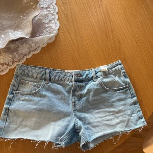 Ljusblå jeansshorts med rå kant - Säljer ett par ljusblå jeansshorts med klassisk femficksdesign och rå, fransig kant nedtill. Shortsen har bälteshällor och knappstängning framtill. De är tillverkade i mjuk bomull och har en avslappnad passform, perfekt för varma dagar. Använt en gång. Storlek L men från ”teens” collection alltså kan vara lite mindre