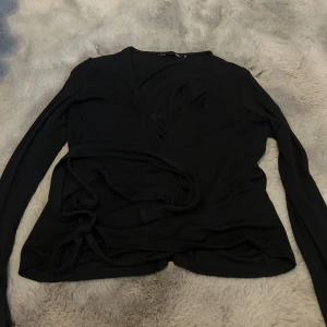 Svart omlottopp med knytband - Svart långärmad omlottopp med v-ringning och knytband i midjan. Snygg och enkel design som passar perfekt till jeans eller kjol. Tunn och mjuk i materialet för en skön känsla.