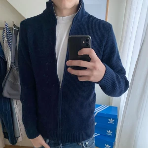 Mörkblå stickad kofta med dragkedja - Säljer en mörkblå stickad kofta med dragkedja och hög krage. Koftan har långärmade ribbade muddar och är perfekt för kyliga dagar. Enkel och stilren design som funkar till jeans eller andra favoritplagg.