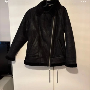 Svart pälsjacka med dragkedja - Snygg svart pälsjacka med bred krage och mjukt foder. Jackan har en sned dragkedja framtill och två fickor på sidorna. Perfekt för kalla dagar. Materialet är pälsimitation och syntet, vilket gör den både varm och stilren.