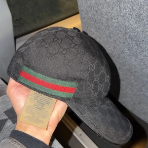 Svart Gucci keps med GG-mönster - Svart keps från Gucci med klassiskt GG-monogram över hela ytan. Kepsen har en böjd skärm och detaljer i grönt och rött på sidan. Tillverkad i bomull med justerbart spänne bak och märkning på insidan. Perfekt för dig som gillar lyxiga streetwear-vibes.