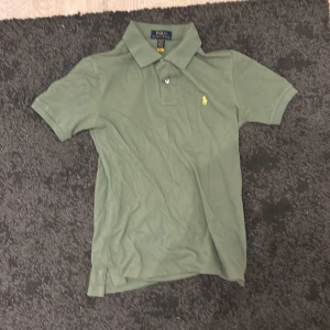 Grön pikétröja från Ralph Lauren - Snygg grön pikétröja från Polo Ralph Lauren med klassisk krage och korta ärmar. Tröjan har den ikoniska gula broderade loggan på bröstet och är gjord i mjuk bomull. Perfekt för dig som gillar stilrena och tidlösa plagg.