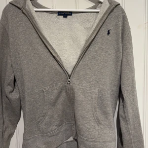 Grå hoodie från Polo Ralph Lauren - Snygg grå hoodie från Polo Ralph Lauren med klassisk blå logga på bröstet. Tröjan har huva, dragkedja. Storleken är dock barnstorlek men i storleken large vilket passar ungefär som S på vanliga tröjor. Hör av er med prisförslag eller funderingar!