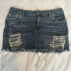 Blå jeanskjol med slitningar Coloring-gem - Trendig blå jeanskjol från Coloring-gem med slitna detaljer och fransig nederkant. Klassisk femficksmodell med dragkedja och knapp framtill. Snyggt trasiga partier framtill ger en cool vibe. Perfekt för dig som gillar en edgy och avslappnad stil. Står ej storlek, jag har S