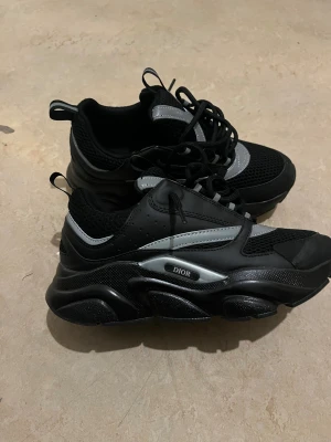 Svarta Dior sneakers med reflexdetaljer - Chunky svarta Dior sneakers med silvriga och vita reflexdetaljer. Ovandel i mesh och skinn, med kraftig sula och snörning. Snygg sportig vibe och tydlig Dior-logga på sidan. Perfekta för dig som gillar statement-sneakers med modern design.