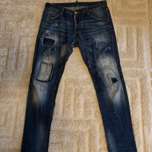 Dsquared2 jeans - Säljer ett par blå dsq2 jeans med coola slitningar och patchade hål på framsidan. Baktill finns två fickor med dragkedjor och en läderdetalj vid midjan. Jeansen har en straight passform och är tillverkade i klassiskt denimtyg.