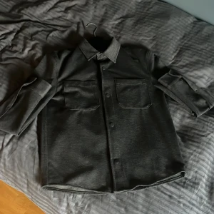 Svart overshirt med fickor - Stilren svart overshirt med två stora bröstfickor och knappar framtill. Skjortan har klassisk krage och långa ärmar som kan vikas upp. Materialet känns robust och har en matt finish, perfekt för lager-på-lager. Enkel och clean design som funkar till många stilar.