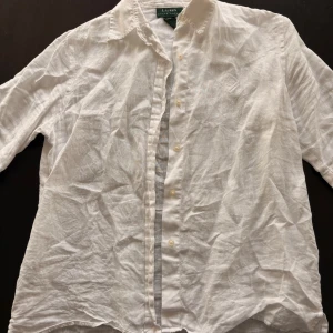 Vit linneskjorta från Ralph Lauren - Vit skjorta i 100% linne från Ralph Lauren, med klassisk krage och knappar framtill. Skjortan har ett snyggt broderat RL-emblem på bröstet och en luftig, normal passform. Perfekt för varma dagar och stilrena looks.