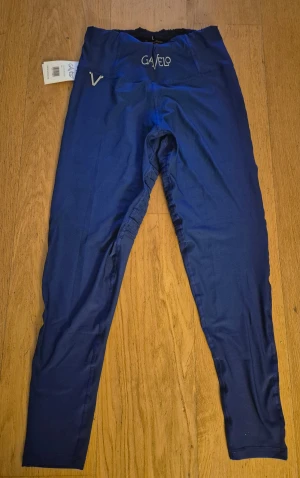 Blå leggings från Gavelo med ribbade detaljer - Ny med Prislappen kvar. Snygga blå leggings från Gavelo med coola ribbade detaljer över knäna och hög midja. Gjorda i stretchigt material av polyester och spandex för perfekt passform. Logga framtill och bak, samt elastisk midja för extra komfort.