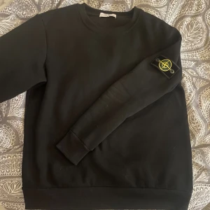 Svart sweatshirt från Stone Island - Svart sweatshirt från Stone Island med klassisk rund hals och ribbade muddar. På vänster ärm sitter den ikoniska Stone Island-patchen med gul broderad kompass. Tröjan är tillverkad i mjuk bomull och har en clean, normal passform som funkar till streetwear.
