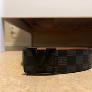 Louis Vuitton rutig svart bälte - Säljer ett stilrent Louis Vuitton bälte med klassiskt rutigt mönster i svart och grått. Bältet har en snygg LV-logga i metall som spänne och är tillverkat i skinn med en slät brun insida. Perfekt accessoar för att lyfta din outfit.