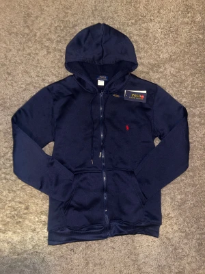 Marinblå hoodie från Polo Ralph Lauren - Snygg marinblå hoodie från Polo Ralph Lauren med dragkedja och huva. Tröjan har ett rött broderat Polo-logo på bröstet, snörning vid huvan och praktiska fickor framtill. Materialet är mjuk bomull som känns skönt mot huden. Perfekt för chill dagar och streetwear.