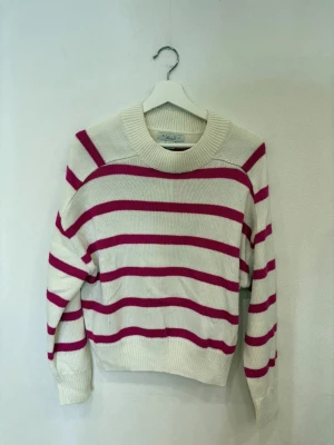 Randig stickad tröja från Amisu - Snygg stickad tröja från Amisu med breda rosa och vita ränder. Modellen har rund hals, ribbade muddar och lång ärm. Perfekt för dig som gillar en chill och färgglad look. Materialet är mjukt och skönt, passar till jeans eller kjol.