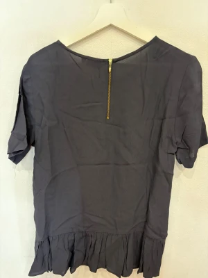 Mörkblå blus med volang från KappAhl - Snygg mörkblå blus från KappAhl i storlek M. Blusen har rund halsringning, korta ärmar och en guldfärgad dragkedja i ryggen. Nedtill finns en bred volang som ger en cool och feminin touch. Perfekt för dig som gillar stilrena detaljer.