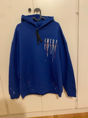 Blå AMIRI hoodie  - Hej säljer denna blå hoodie från AMIRI med färgstänksmönster och stor logga på ryggen. Tröjan har huva och långa ärmar, samt tryck på bröstet och ryggen. Perfekt för dig som gillar streetwear och vill sticka ut. Storlek M men sitter större så passar även L.