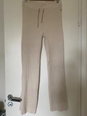 Beige ribbade mysbyxor från Pernilla Wahlgrens design - Snygg beige ribbade mysbyxor från Pernilla Wahlgren Design i storlek L. Byxorna har en mjuk stickad känsla, snörning i midjan, bred resår och en liten guldfärgad detalj med ananas. Perfekt för dig som gillar stilrena och bekväma plagg med twist. Byxorna är aldrig använda. Säljes pga felköp.