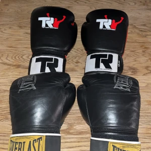 Svarta boxningshandskar Top Rank & Everlast - Två par svarta boxningshandskar, ett från Top Rank Equipment med vit och röd logga och ett från Everlast med gul och vit logga. Båda har klassisk form och bred kardborre vid handleden. Perfekt för träning och sparring.