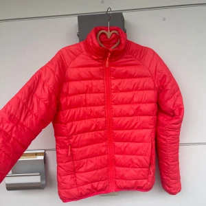 Röd pufferjacka med dragkedja - Säljer en koral pufferjacka med horisontella quiltade paneler och hög krage. Jackan har två sidofickor med dragkedja och hel dragkedja framtill. Materialet är syntet och jackan är långärmad, perfekt för kyliga dagar.Höst/vår jacka