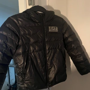 Svart pufferjacka från EA7 Armani - Säljer en svart pufferjacka från EA7 Emporio Armani med huva och två sidofickor med dragkedja. Jackan har ett stilrent EA7-märke på bröstet och quiltad design som ger en cool look. Perfekt för kalla dagar och enkel att matcha med din outfit. För barn Stl 130