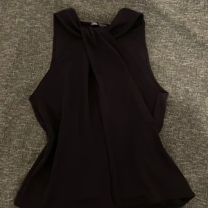 Svart halterneck topp från Zara - Snygg svart halterneck från Zara med twistad detalj vid halsen. Toppen är ärmlös och har en lösare passform. Säljer då den inte kommer till användning