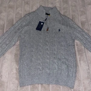 Grå stickad tröja Polo Ralph Lauren - Snygg grå kabelstickad tröja från Polo Ralph Lauren med half zip och hög krage. Tröjan har långärmade ribbade muddar och diskret broderad logga på bröstet. Perfekt för dig som gillar klassisk stil med en modern touch.