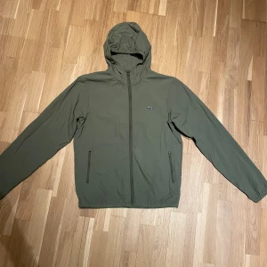 Olivgrön vindjacka från Lacoste - Snygg olivgrön vindjacka från Lacoste med huva och hel dragkedja framtill. Jackan har två sidofickor med dragkedja och den klassiska krokodilloggan på bröstet. Perfekt för dig som gillar stilren och sportig look.