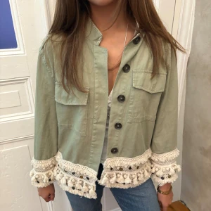 Grön jeansjacka med virkade detaljer - Snygg ljusgrön overshirt med stora svarta knappar och två bröstfickor. Jackan har unika virkade detaljer i beige med tofsar och pärlor längs nederkant och ärmslut. Perfekt för dig som gillar boho-vibe och vill sticka ut.