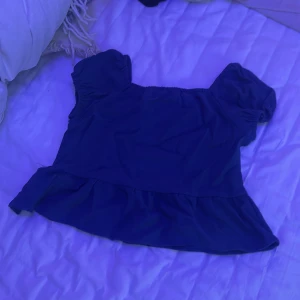 Blå volangtopp med puffärm - Supersöt blå/ mörkblå topp med volang nertill och puffiga korta ärmar. Toppen har en fyrkantig ringning och är gjord i ett lätt, strukturerat bomullstyg som känns luftigt. Perfekt för dig som gillar en feminin och trendig stil.