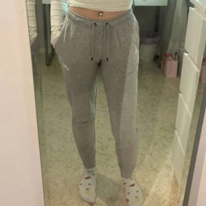 Grå mjukisbyxor från Nike med snörning - Grå mjukisbyxor från Nike med klassisk logga på vänster sida. Byxorna har resår i midjan, snörning och muddar vid bensluten. Tillverkade i mjukt bomullsmaterial, perfekt för chill eller träning. Snygg och enkel design som är lätt att matcha.