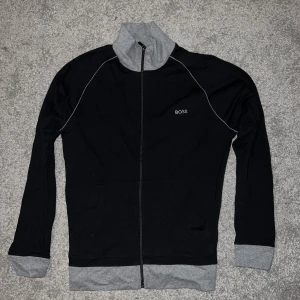 Svart ziptröja från BOSS med grå detaljer - Svart långärmad ziptröja från BOSS med grå krage, muddar och nederkant. Tröjan har hel dragkedja, fickor framtill och vit piping längs axlarna. Snygg och enkel design med BOSS-logga på bröstet.