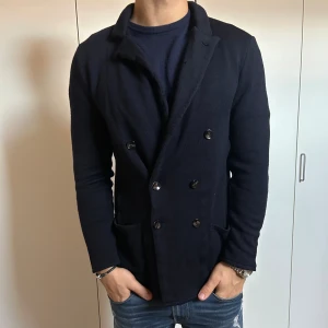 Mörkblå stickad kavaj med knappar - Snygg mörkblå stickad kavaj med dubbelknäppning och två framfickor. Kavajen har en klassisk krage och långärmade ärmar, perfekt för en chill och stilren look. Passformen är normal och materialet känns mjukt och bekvämt mot kroppen.