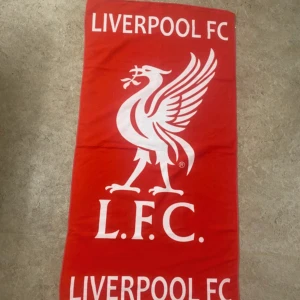 Liverpool FC röd halsduk - Röd halsduk med stort vitt Liverpool FC-tryck och klubbens klassiska fågelmotiv. Perfekt accessoar för dig som vill visa ditt stöd på matchen eller i vardagen. Halsduken har en rektangulär form och tydliga klubbnamn i båda ändar.