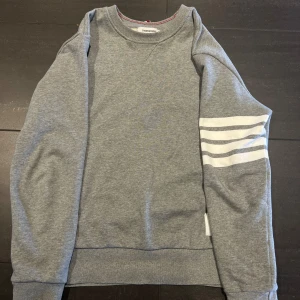 Grå sweatshirt från Thom Browne - Säljer en grå sweatshirt från Thom Browne med klassiska vita ränder på ena ärmen och ribbade muddar. Tröjan har rund halsringning och är tillverkad i mjuk bomull. Perfekt för en clean och stilren look. Pris kan diskuteras