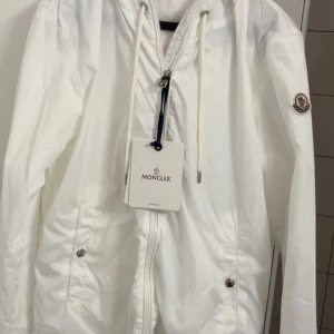 Vit vindjacka från Moncler - Snygg vit vindjacka från Moncler med huva och snörning. Jackan har dragkedja framtill, två fickor med tryckknappar och Moncler-logga på ärmen. Perfekt för dig som vill ha en clean och stilren look.