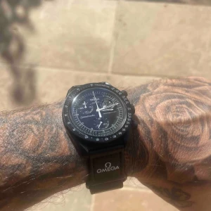 Omega swatch snoppy  - Säljer en stilren svart Omega swatch Speedmaster med rund boett och tachymeter-skala. Klockan har svart urtavla med vita detaljer och tre subdials. Armbandet är svart med Omega-logga. Perfekt för dig som gillar klassisk design och ikoniska märken. Öppen för byten med. 