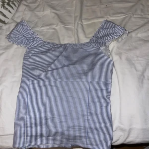 Blårandig blus från Subdued - Säljer en söt blus från Subdued i storlek M. Blusen är blårandig med vita knappar framtill, har offshoulder-ärmar med volangkant. Materialet är mjukt och känns som bomull.