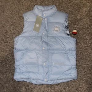 Ljusblå dunväst från Canada Goose - Snygg ljusblå dunväst från Canada Goose med hög krage och tryckknappar framtill. Västen har två stora fickor med logotyp och ett broderat märke på bröstet. Insidan har ett coolt mönster i blått och vitt. Perfekt för kalla dagar när du vill se chill ut.