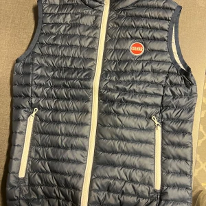 Mörkblå dunväst från Colmar - Snygg mörkblå dunväst från Colmar med quiltad design och vita dragkedjor. Västen har två fickor med dragkedja och ett broderat Colmar-märke på bröstet. Perfekt att slänga över en hoodie för extra värme. Den är i skick: 10/10, precis som ny! Det är bara att skriva vid minsta fråga, priset är inte hugget i sten!